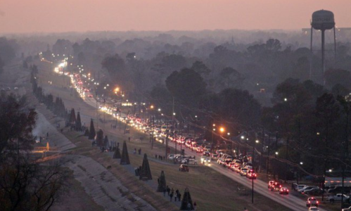 Aerial view of a long line of cars on a road at dusk, with trees and a water tower in the background.