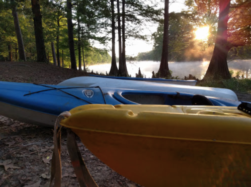 Two kayaks, one blue and one yellow, are resting on a sandy shore with a misty river and trees in the background at sunrise.