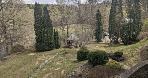 A serene garden scene featuring a gazebo surrounded by trees and shrubs, with a grassy area and a distant car.