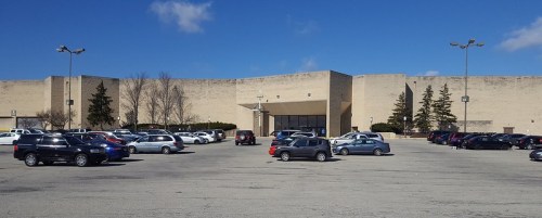 A large, empty parking lot in front of a beige shopping mall under a clear blue sky.