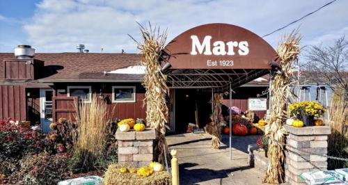 A rustic storefront with a sign reading "Mars, Est 1923," decorated with pumpkins and autumn plants.