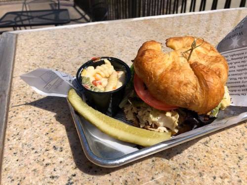 A croissant sandwich with lettuce, tomato, and meat, served with macaroni salad and a pickle on a tray.