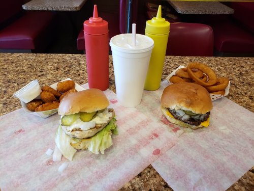 Two burgers with toppings, a side of onion rings, and dipping sauce, alongside three soft drink cups in a diner setting.