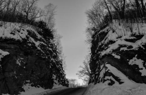 A snow-covered road between steep, rocky cliffs, with bare trees silhouetted against a gray sky.