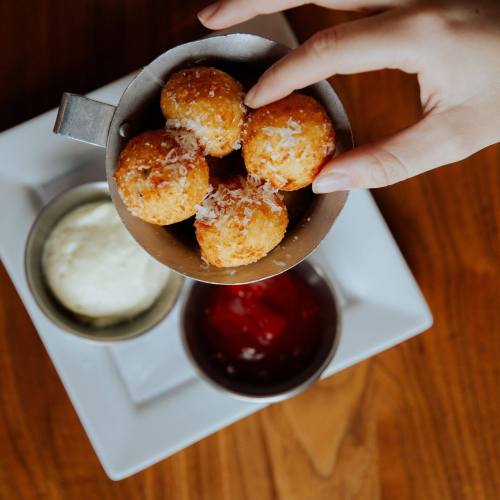 A hand holding a small bowl of golden, cheesy balls, accompanied by dipping sauces on a wooden table.