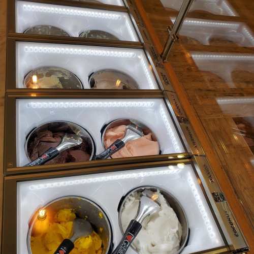 A display of various gelato flavors in metal tubs, illuminated with bright lights.