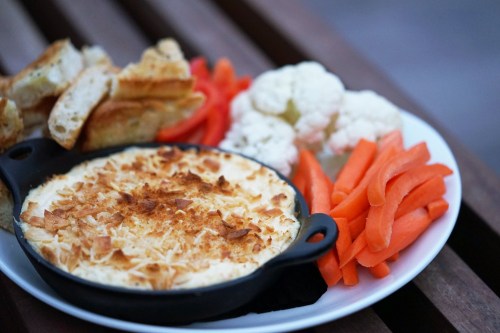 A plate with baked dip, toasted bread, carrot sticks, and cauliflower florets on a wooden surface.