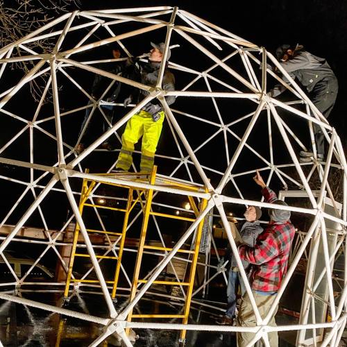 A group of people assembling a large geodesic dome at night, using ladders and tools.