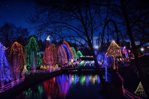 A festive scene with colorful lights adorning trees and structures by a calm river at night.