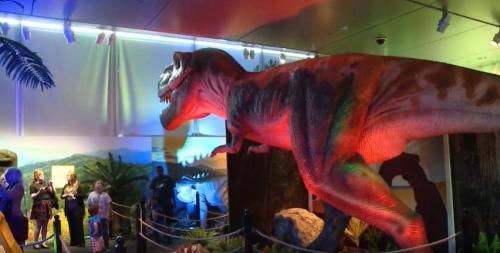 A life-sized dinosaur exhibit with a T. rex model, colorful lighting, and visitors admiring the display.