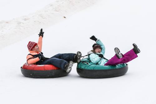 Two people in colorful winter attire joyfully tubing down a snowy slope, raising their arms in excitement.