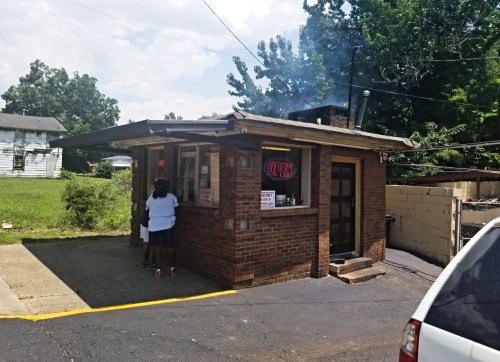 A small brick building with an "OPEN" sign, surrounded by greenery, with two people at the counter.