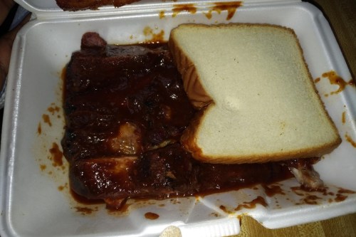 A takeout container with barbecue ribs covered in sauce and a slice of bread on top.