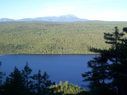 A serene landscape featuring a blue lake surrounded by dense green forests and distant mountains under a clear sky.