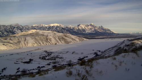 Snow-covered mountains rise above a serene valley, with fields and a river partially frozen in the foreground.