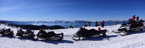 A panoramic view of snowmobiles on a snowy mountain, with people enjoying the scenic lake and mountain backdrop.