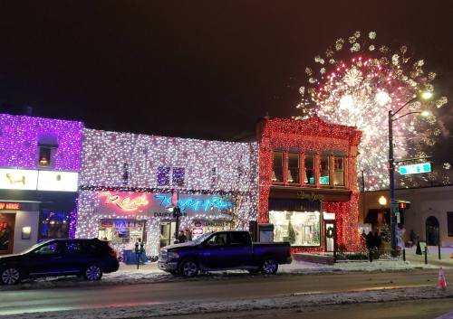 Colorful holiday lights adorn buildings, with fireworks bursting in the night sky above a snowy street scene.