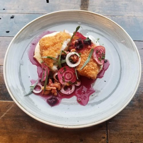 A plated dish featuring crispy fish, creamy sauce, pickled vegetables, and a vibrant berry sauce on a rustic wooden table.