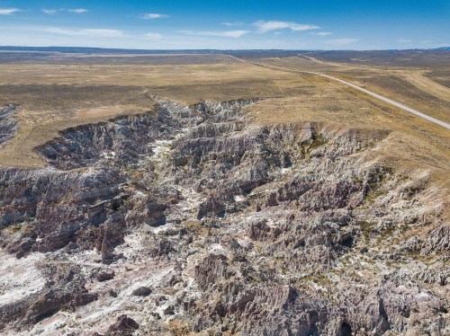 Aerial view of a rugged, colorful canyon landscape with dry terrain and a distant road under a clear blue sky.