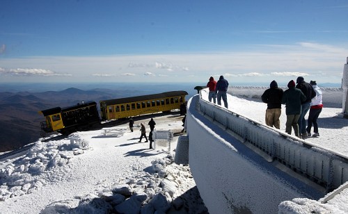A yellow train at a snowy mountain summit, with people exploring the area and enjoying the scenic view.