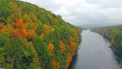A scenic river flows through vibrant autumn foliage, with trees displaying shades of orange, yellow, and green under a cloudy sky.