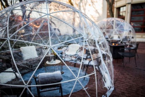Two transparent geodesic domes set up outdoors, featuring cozy seating and blankets, surrounded by a brick patio.