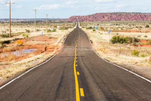 A long, straight road stretches into the distance, flanked by dry grasslands and red rock formations under a blue sky.