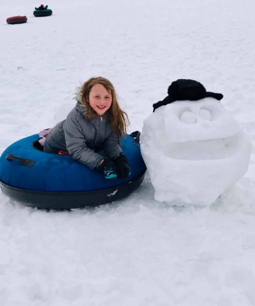 A smiling girl sits on a blue snow tube next to a snowman with a hat, on a snowy landscape.