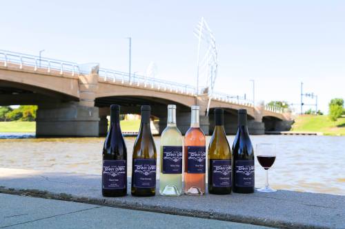 Bottles of wine arranged by a river with a bridge in the background, featuring red, white, and rosé varieties.