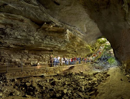 A group of people walking along a path inside a large cave, with rocky walls and a bright opening in the distance.