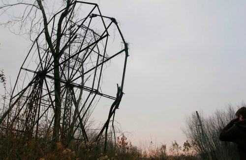 A person stands near a dilapidated ferris wheel, overgrown with vines, against a gray sky.