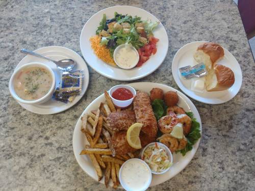 A table with a meal featuring fried fish, fries, shrimp, coleslaw, salad, soup, and rolls with butter.