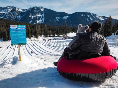 A person sits on a snow tube with a child, overlooking a snowy tubing hill and mountains in the background.