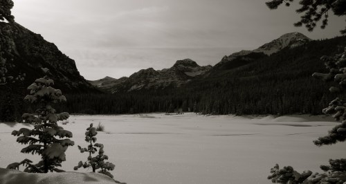 A serene winter landscape featuring snow-covered mountains and a frozen lake, framed by evergreen trees.
