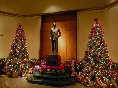 A statue of a man stands between two decorated Christmas trees, surrounded by gifts and festive decorations.