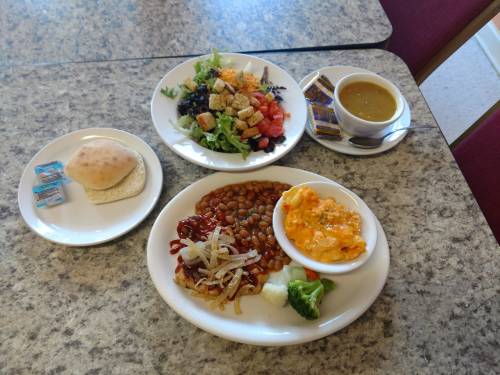 A plate with baked beans, macaroni and cheese, broccoli, and onions, alongside a salad, bread roll, and soup.