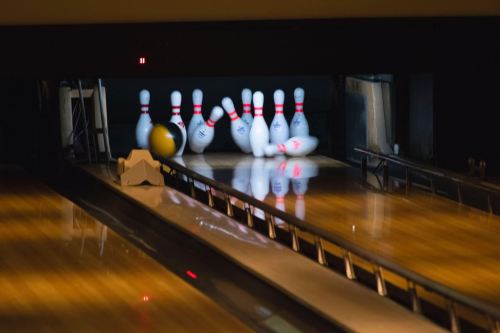 A bowling ball rolls down the lane, striking pins and causing some to fall, with reflections visible on the polished wood.