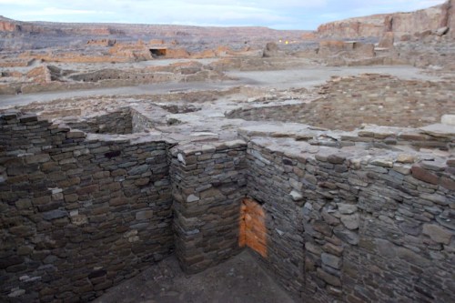 Ancient stone ruins in a desert landscape, with rocky terrain and distant cliffs under a cloudy sky.
