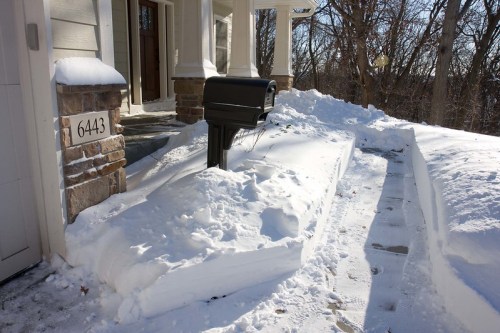 A mailbox surrounded by deep snow, with a clear path leading to the front door of a house.