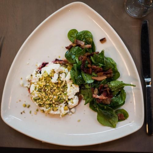 A triangular plate with ricotta topped with pistachios and a spinach salad with bacon bits on the side.