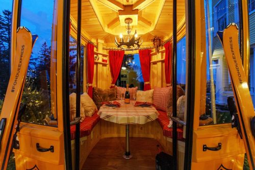 Cozy dining space with wooden decor, red curtains, and a chandelier, set for fondue, illuminated at dusk.