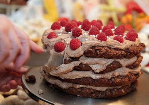 A hand serves a layered chocolate cake topped with raspberries and silver sprinkles on a decorative platter.