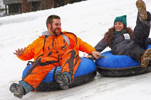 Two people in winter gear joyfully tubing down a snowy slope, with snowflakes falling around them.