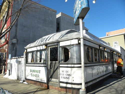 A vintage diner with a white exterior and a sign reading "Summit Diner," surrounded by snow and blue sky.