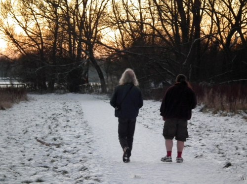 Two people walk along a snowy path at sunset, surrounded by trees and a serene winter landscape.