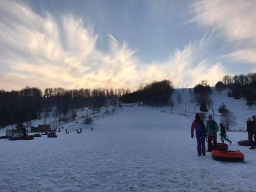 A snowy hill with people tubing, surrounded by trees and a colorful sky at sunset.