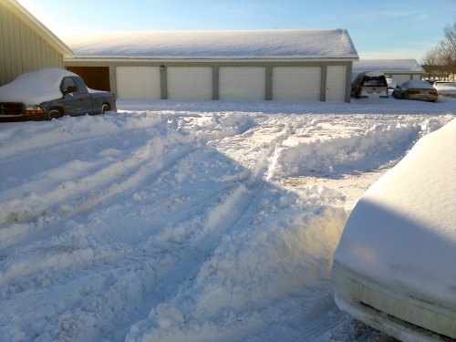A snowy scene showing tire tracks in deep snow, with parked cars and a garage in the background under a clear blue sky.