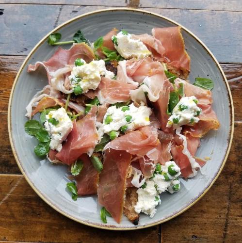 A plate of prosciutto, fresh peas, and creamy ricotta on toasted bread, garnished with greens.