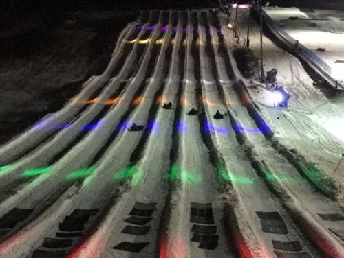 Aerial view of a snow tubing slope at night, illuminated with colorful lights and featuring several people sliding down.