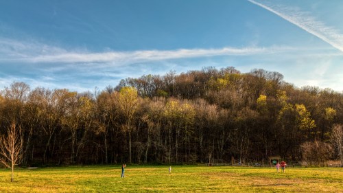 A grassy field with three people in the foreground and a wooded hill in the background under a blue sky.